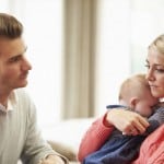 Anxious mother consumed by worry holds baby while staring blindly into the distance. Supportive husband looks upon his wife with concern.