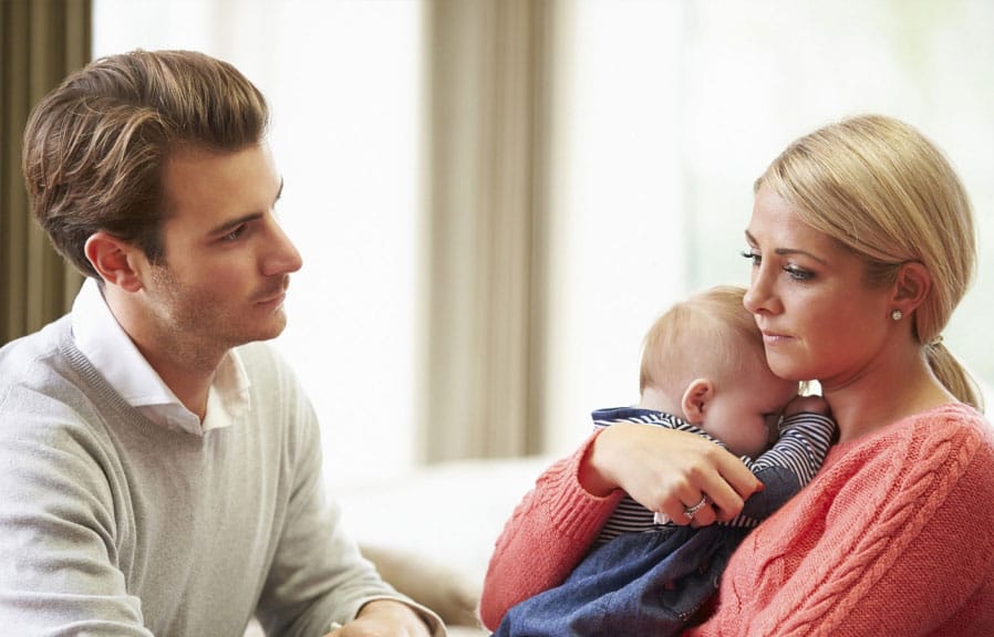 Anxious mother consumed by worry holds baby while staring blindly into the distance. Supportive husband looks upon his wife with concern.