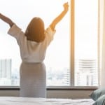 A well rested woman stretches in front of a window at sunrise