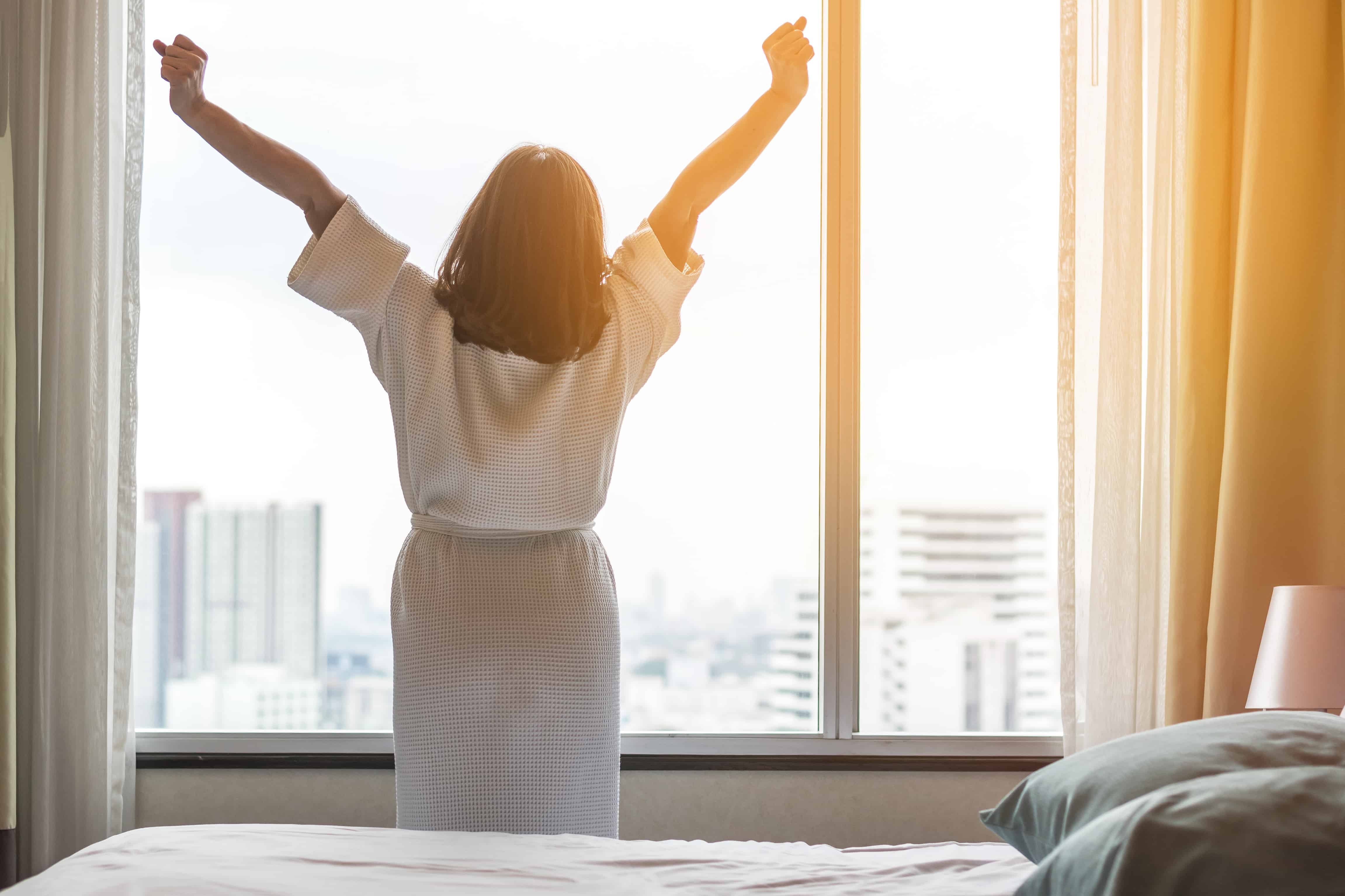 A well rested woman stretches in front of a window at sunrise