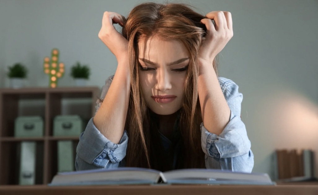 College student studying at a desk while feeling overwhelmed, holding her head in stress while looking at an open book.