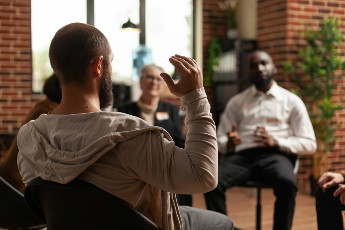 Man sits with raised, animated hands, one among a circle of addicts sharing in a group counseling session.