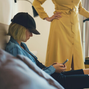 Rebellious drug addicted teen wearing black had and blue jacket looks downward at her phone ignoring her concerned mother.