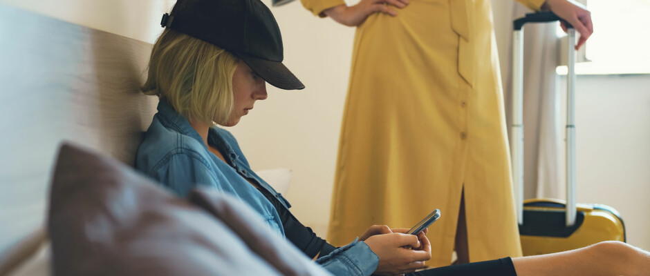 Rebellious drug addicted teen wearing black had and blue jacket looks downward at her phone ignoring her concerned mother.