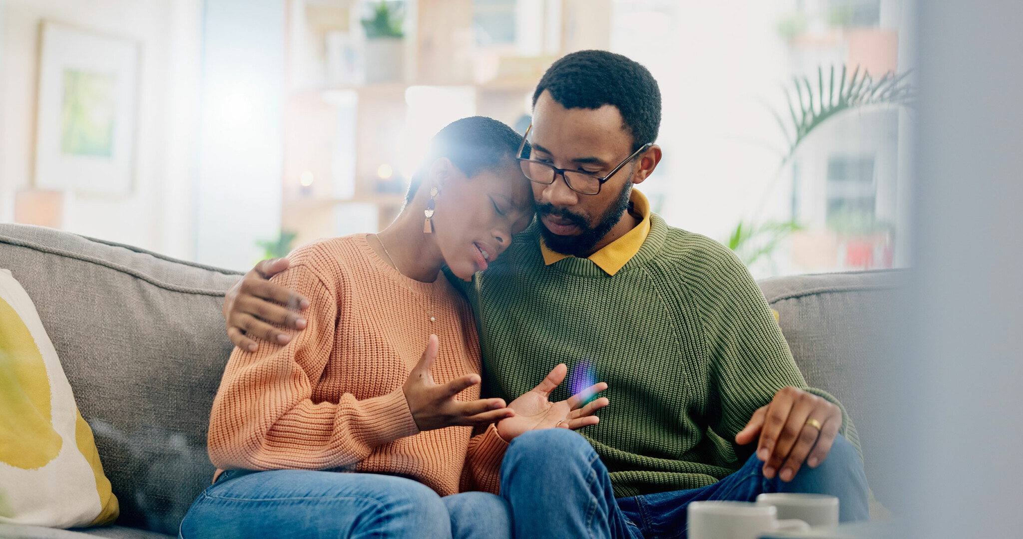 Black couple sitting on couch in an emotional embrace. Anxiety-ridden woman speaks with palms facing upwards while attentive husband provides understanding and support.