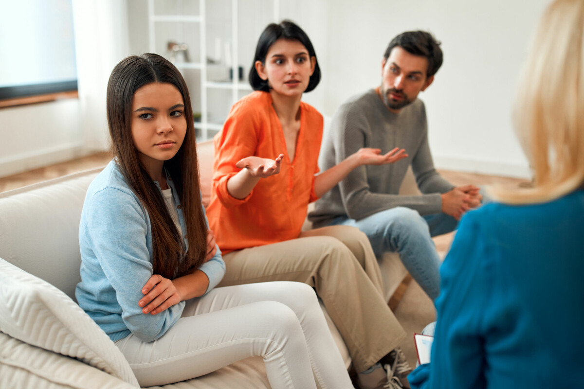 Frowning and staring into the distance, an upset teenage girl sits alongside exasperated parents at a family counseling appointment.