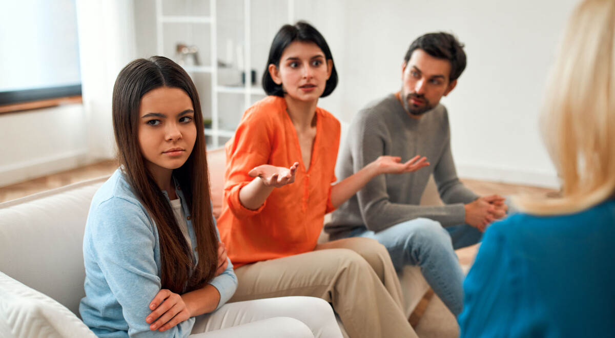 Frowning and staring into the distance, an upset teenage girl sits alongside exasperated parents at a family counseling appointment.
