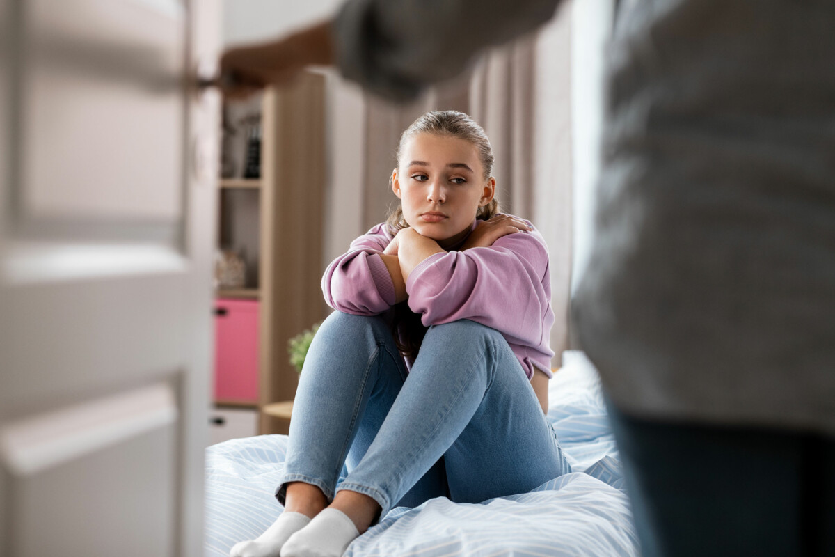 Mother opens door to reveal frustrated daughter sitting on bed at home.