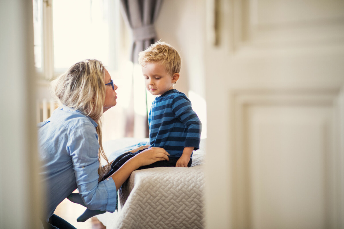 A young mother talks to her toddler son inside in a bedroom.