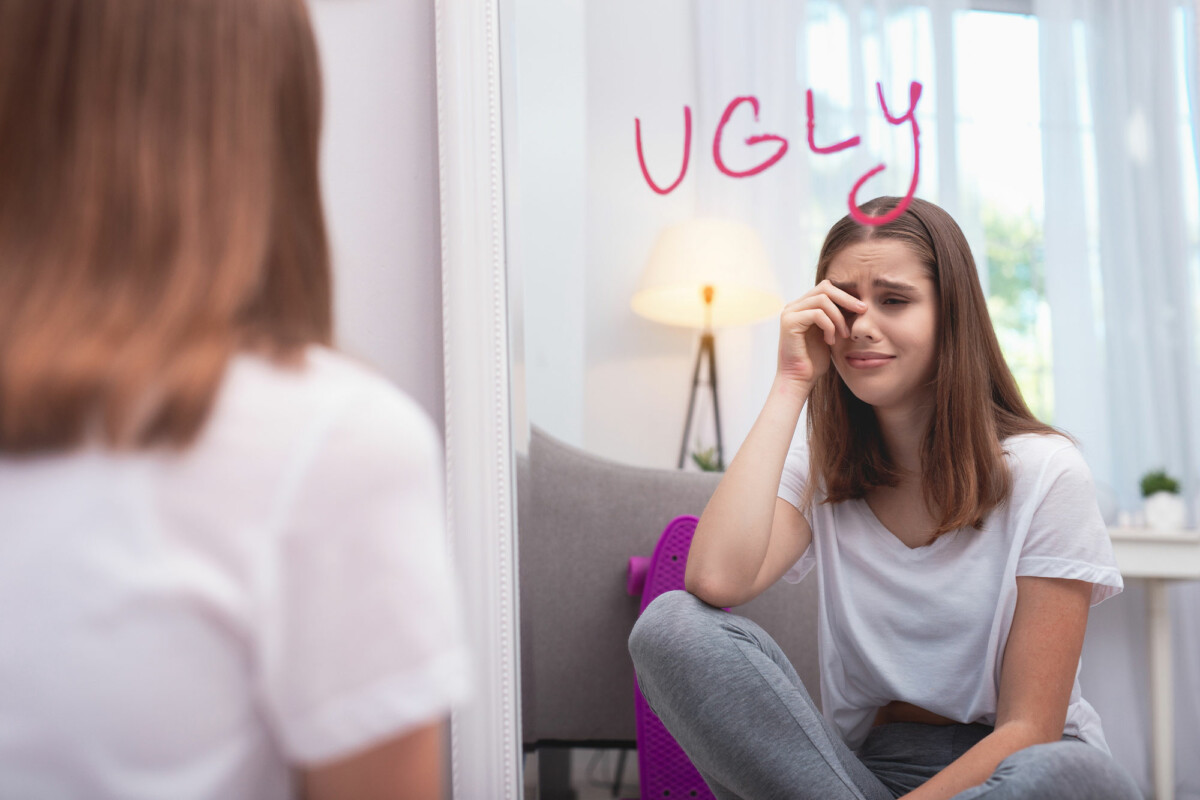 Crying teen girl looks into mirror with the word ugly written on it in lipstick