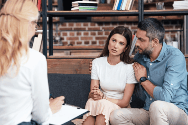 A female psychiatrist takes notes as a couple embraces during a therapy session.