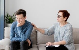 Woman with short red hair and glasses comforts a man sitting on a sofa, placing a reassuring hand on his shoulder as he looks down, in a living room.