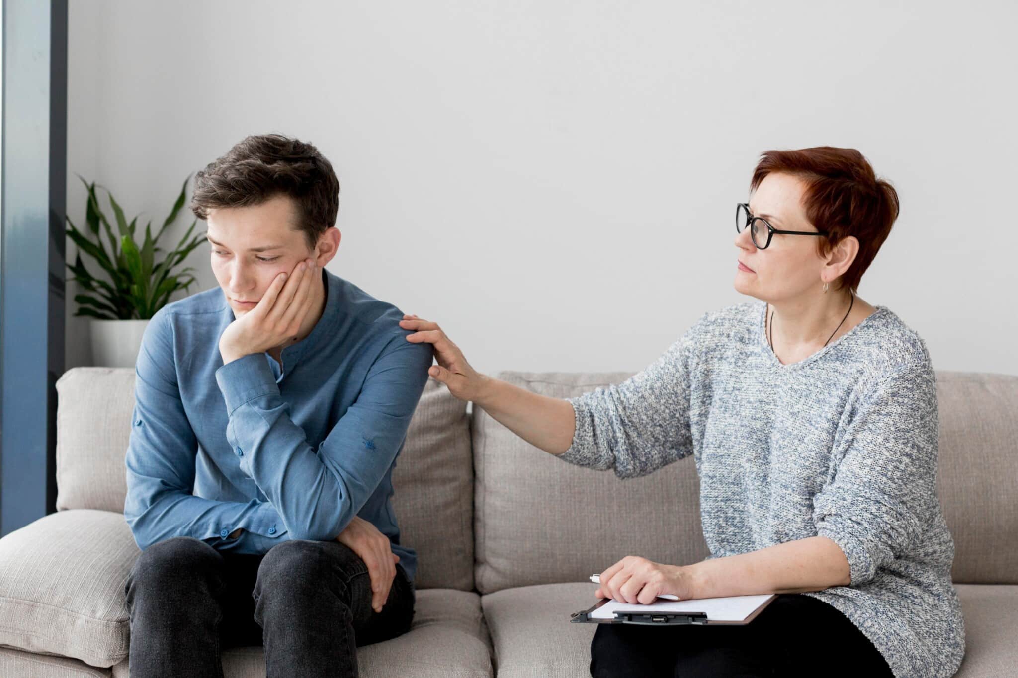 Woman with short red hair and glasses comforts a man sitting on a sofa, placing a reassuring hand on his shoulder as he looks down, in a living room.
