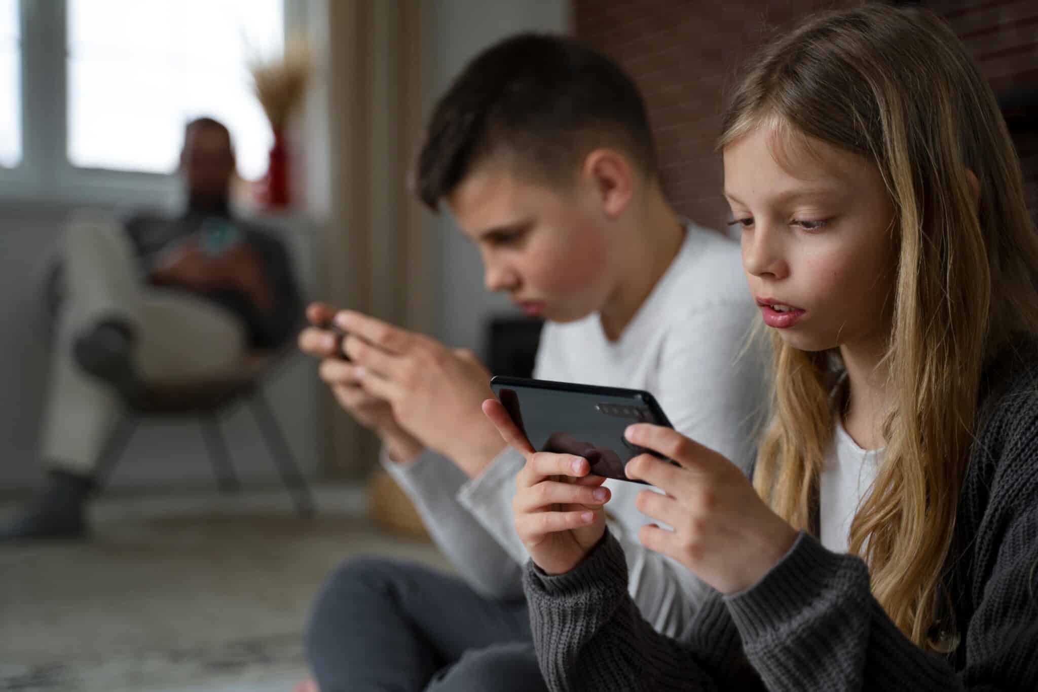 Two children sit closely indoors, each focused on a handheld gaming console.
