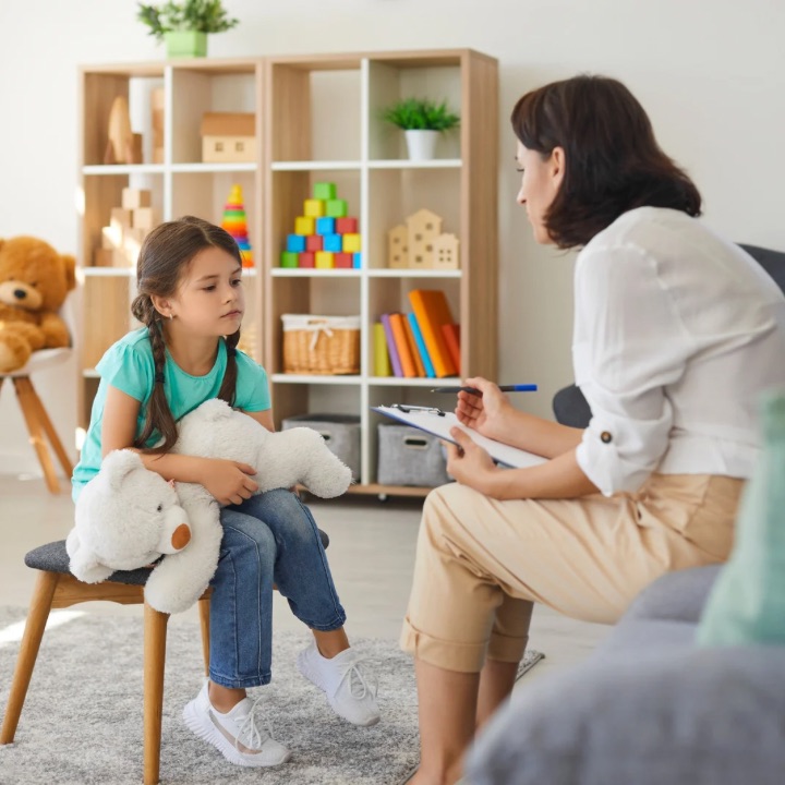image -Child sitting on a chair holding a teddy bear, while a psychologist conducts an evaluation. 2025-10-17T115416702 Child sitting on a chair holding a teddy bear, while a psychologist conducts an evaluation.