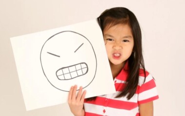 Young girl holding a drawing of an angry face while making an angry expression.