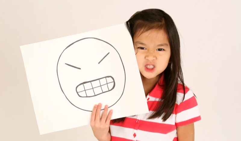 Young girl holding a drawing of an angry face while making an angry expression.