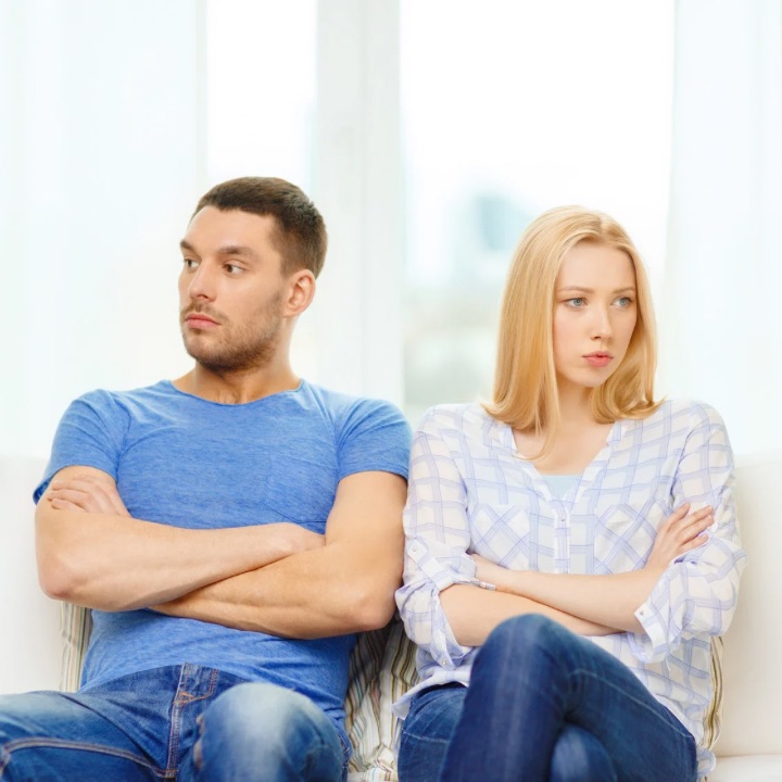 Couple sitting on a couch with arms crossed, appearing upset and distant from each other.