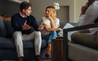 A couple sitting on a couch having a serious discussion during a couples therapy session with a therapist taking notes.