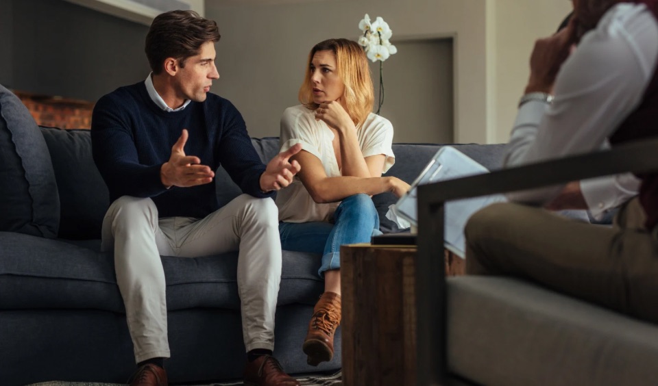 A couple sitting on a couch having a serious discussion during a couples therapy session with a therapist taking notes.