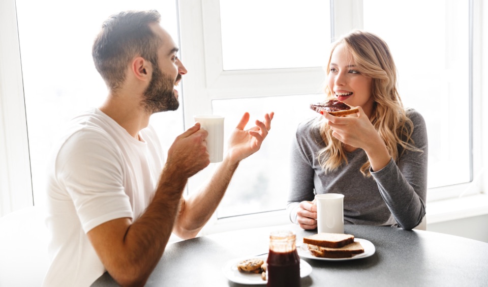 A smiling couple sitting at a breakfast table, enjoying coffee and toast while having a relaxed conversation near a bright window.