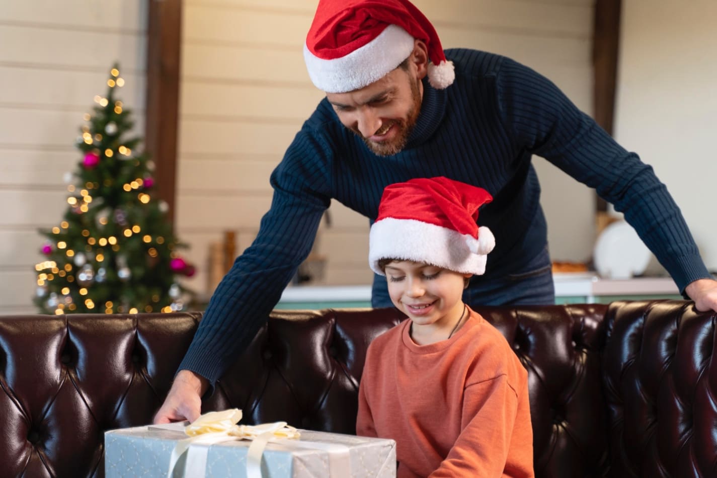 Dad wearing a Santa hat helps his smiling child open a wrapped Christmas gift while sitting together on a couch with a decorated tree in the background. - Total Life Counseling for Children, Teens, Adolescents and Adults in Orlando Winter Park Clermont Lake Mary and Central Florida Dad wearing a Santa hat helps his smiling child open a wrapped Christmas gift while sitting together on a couch with a decorated tree in the background.