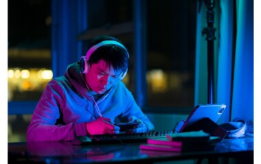 Person wearing headphones at a desk under blue-pink neon lighting, focused on a handheld device with a keyboard nearby