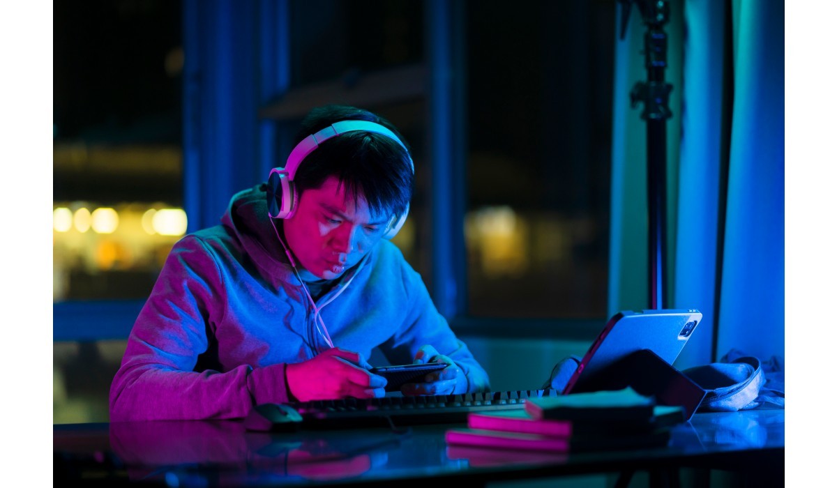 Person wearing headphones at a desk under blue-pink neon lighting, focused on a handheld device with a keyboard nearby