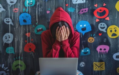 Young person in a red hoodie sits with head in hands over a laptop, surrounded by colorful emoji graffiti on a dark wall, conveying online distress or cyberbullying.
