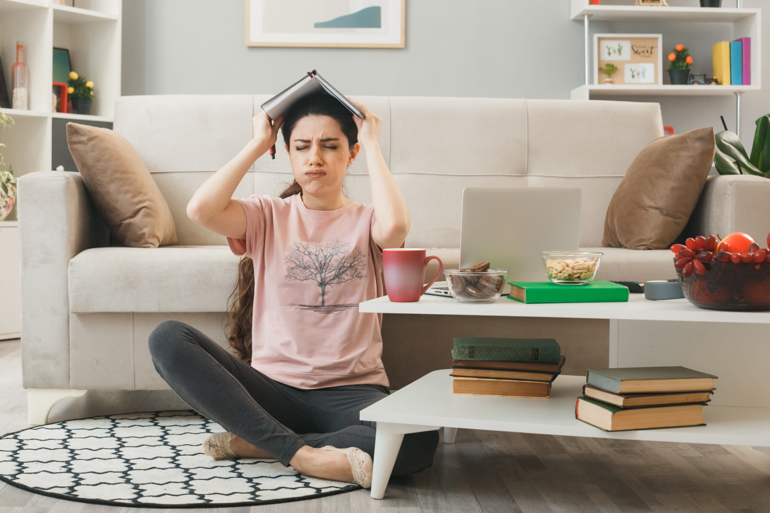 Woman sitting cross-legged on a rug, balancing a notebook on her head with a puzzled expression at a coffee table full of snacks and books in a living room.