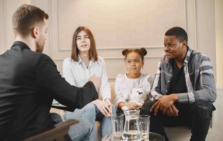 Four people seated in a living room, engaged in a discussion around a glass coffee table with drinks.