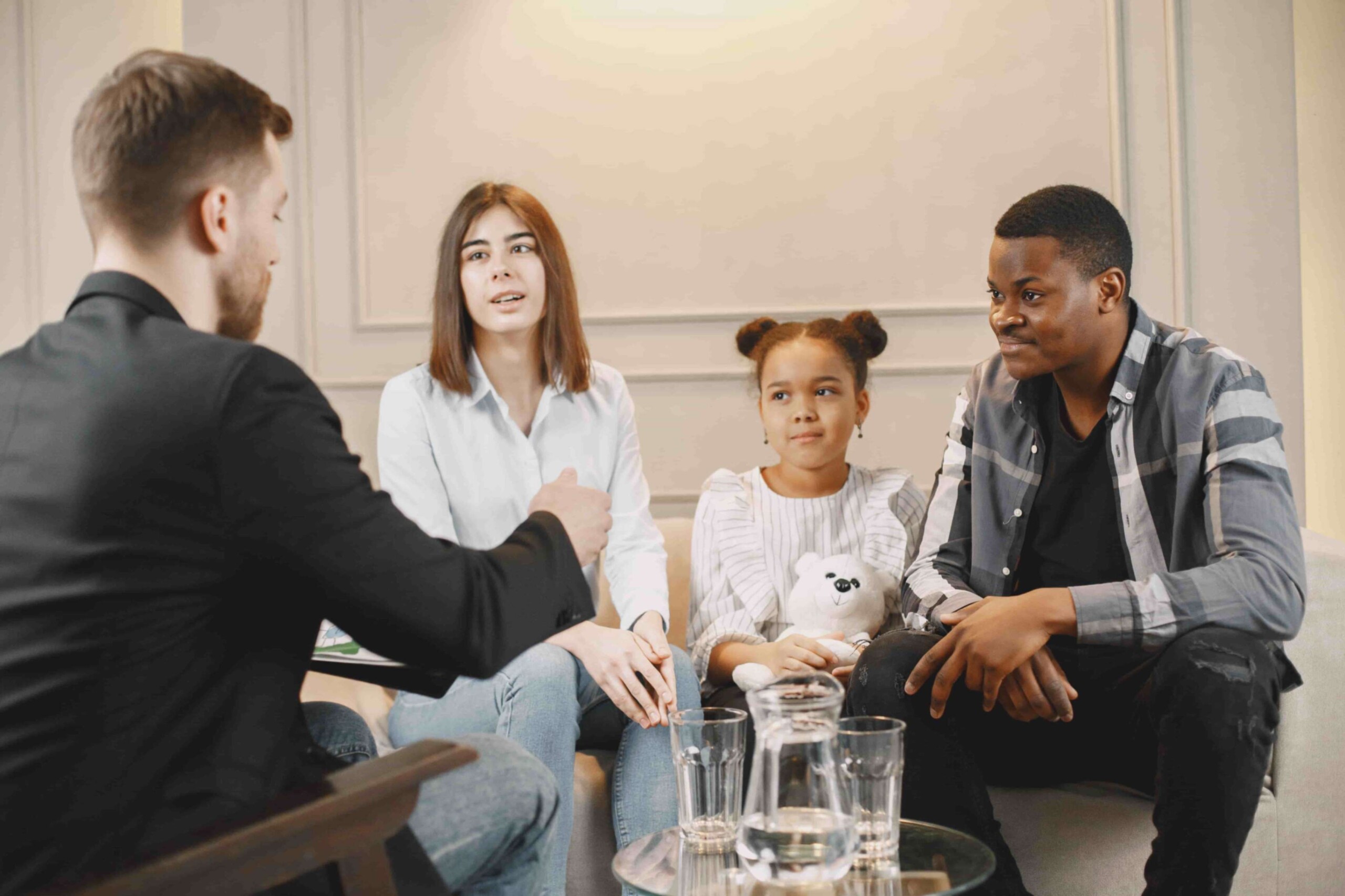 Four people seated in a living room, engaged in a discussion around a glass coffee table with drinks.